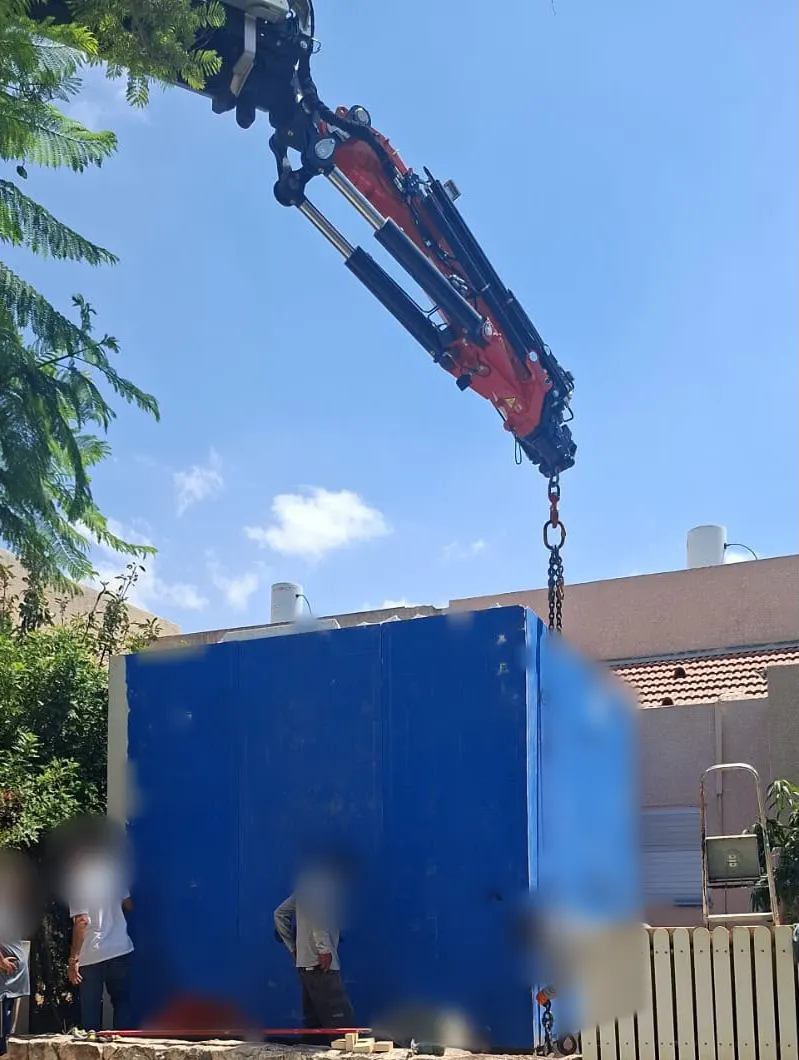 מעטפת ממ"ד תלויה במנוף מעל חצר של בית פרטי במכבים רעות, רגע לפני ההצבה במקומה
