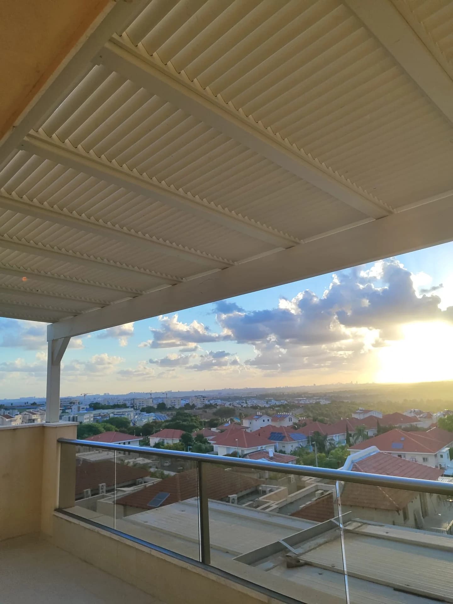 פרגולת אלומיניום לבנה עם רפפות אורך, מרפסת דירה גבוהה עם נוף לעיר ושקיעה