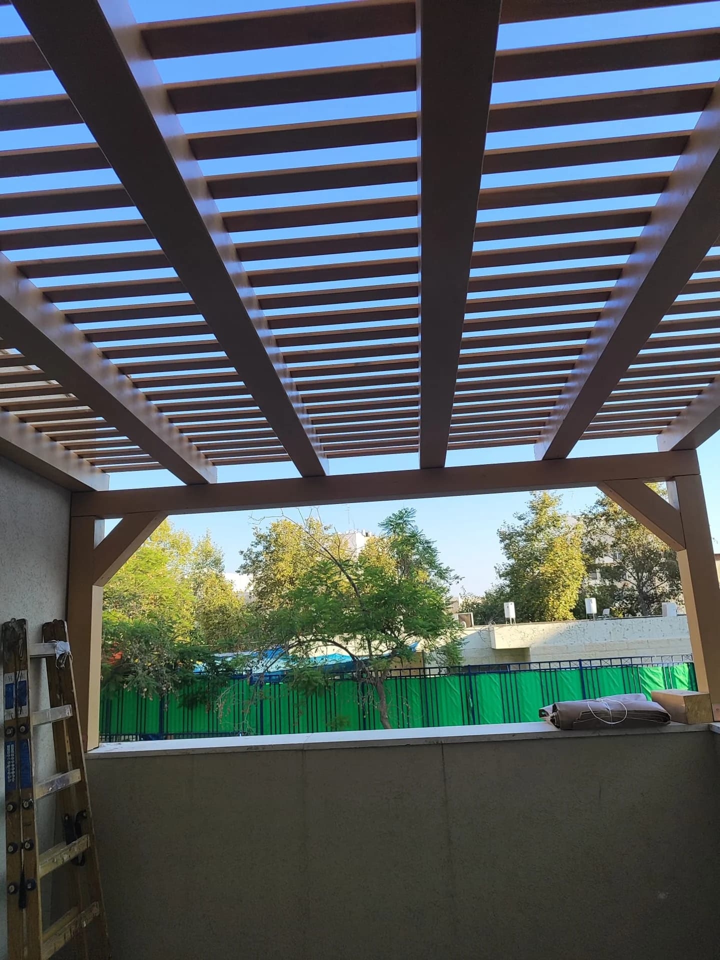פרגולת עץ בגוון חום-אדום על מרפסת בית פרטי, פתחים לכיוון נוף עם גדר ירוקה