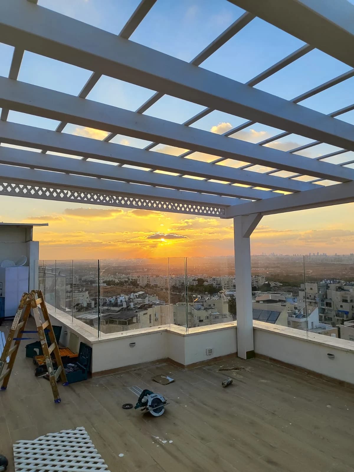 פרגולת עץ לבנה עם קורות אופקיות ומעקה זכוכית, נוף לעיר ושקיעה
